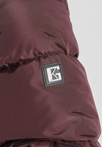 Burgundowa kurtka puchowa z pikowanym wzorem, z czarną prostokątną naszywką z logo "KG" w kolorze srebrnym. Gładki, błyszczący materiał.