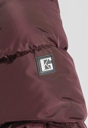 Sección de chaqueta acolchada burdeos con textura acolchada, que presenta un parche rectangular negro con el logotipo plateado "KG". Tejido suave y brillante.