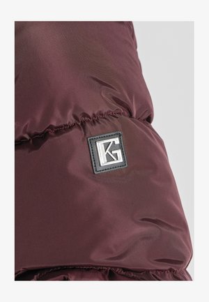 Sección de chaqueta acolchada burdeos con textura acolchada, que presenta un parche rectangular negro con el logotipo plateado "KG". Tejido suave y brillante.