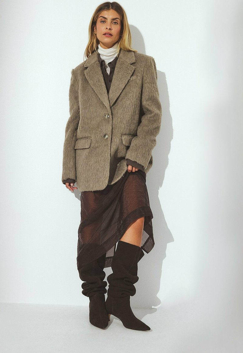 Blazer oversize marrone e textured con due bottoni, indossato sopra una gonna scura semitrasparente e stivali marroni al ginocchio con effetto slouch.