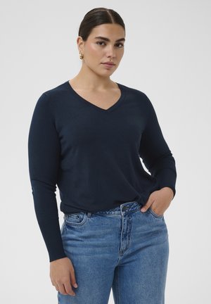 Sötét hajú nő, tengerészkék hosszú ujjú V-nyakú pulóverben és kék farmerben, egy kézzel a zsebében áll egy egyszerű háttér előtt.