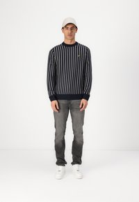 Marine gestreifter Pullover mit weißen vertikalen Linien, mit gerippten Bündchen und Saum. Kombiniert mit grauen Jeans und weißen Sneakern. Militärische Kappe in Weiß.