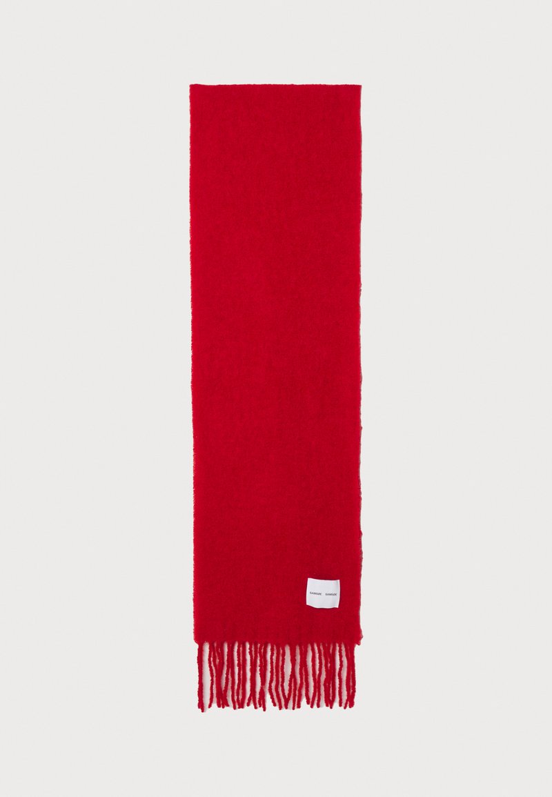 Écharpe rouge en tissu doux et texturé. Présente de longues fringes à chaque extrémité et une petite étiquette de marque blanche cousue sur un côté.