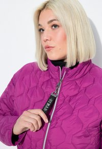 Žena s blond bob �účesem, oblečená v fialové prošívané bundě, drží táhlo zipu s nápisem "HYPRAR" blízko brady.