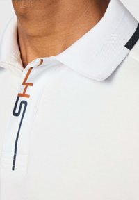 Detailný záber osoby v bielej polo tričku s textúrovaným golierom a modro-oranžovou grafickou potlačou pozdĺž gombíkového zapínania.