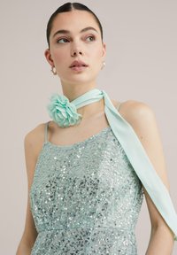 Ljus teal klänning med paljetter, spaghetti-bandsaxlar och en sidentu szak med en tygblomma som dekoration vid halsen.