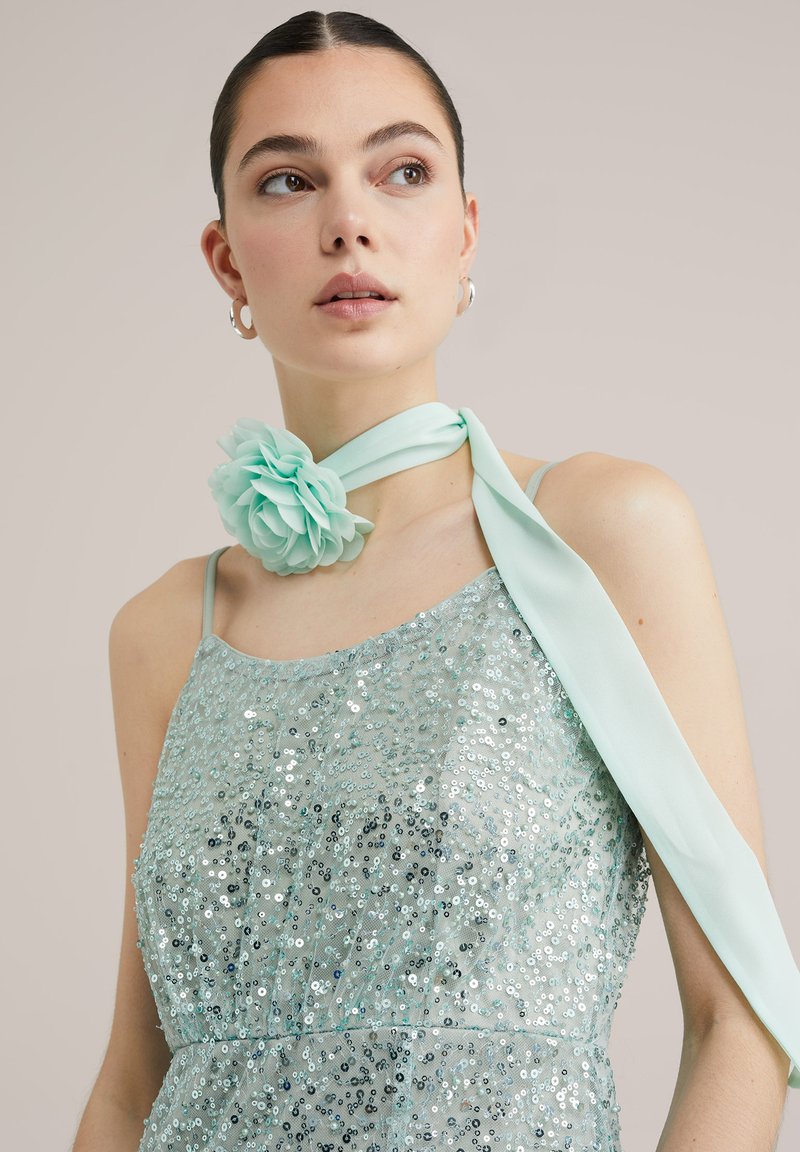 Ljus teal klänning med paljetter, spaghetti-bandsaxlar och en sidentu szak med en tygblomma som dekoration vid halsen.