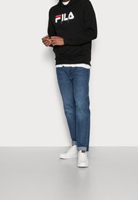 Man i svart FILA-hoodie, vit T-shirt, blå uppkavlade jeans och vita sneakers som står mot en enfärgad ljus bakgrund.