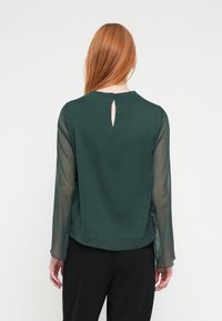 Zelená blúzka s priesvitnými dlhými rukávmi, zaobleným výstrihom a kľúčovým otvorom na chrbte. Hladká textúra a pohodlný strih.