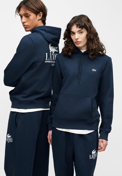 Два модела носят идентични тъмносини суичъри и спортни панталони на Lacoste; единият е обърнат напред, а другият показва логото на Lacoste на гърба.