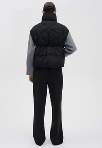 Schwarze gepolsterte Weste mit hohem Kragen, die ein gestepptes Muster aufweist und einen leicht verkürzten Schnitt hat. Kombiniert mit weit geschnittenen schwarzen Hosen.