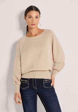Femme aux cheveux foncés vêtue d'un pull beige et d'un jean bleu foncé, debout devant un fond clair, portant des boucles d'oreilles pendantes et une main dans la poche.