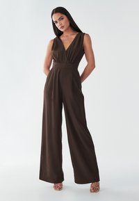 Smeđa kombinezon širokih nogavica s dubokim V-izrezom i bez rukava. Izrađen od glatkog materijala, ima pristajajući struk i naborane bočne stranice.
