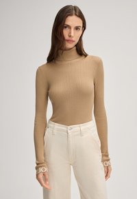 Hellbrauner gerippter Rollkragenpullover mit langen Ärmeln, der kontrastierende weiße Logo-Akzente an den Bündchen aufweist, kombiniert mit weißen Hosen.