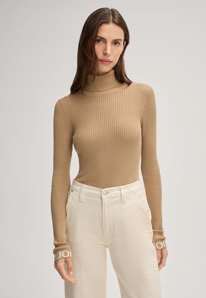 Maglione a collo alto ribbed marrone chiaro con maniche lunghe, con accenti a logo bianco a contrasto sui polsini, abbinato a pantaloni bianchi.