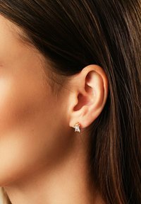 Nahaufnahme eines Frauenohres mit glattem braunen Haar, das einen kleinen goldenen Stecker mit einem klaren rechteckigen Edelstein trägt.