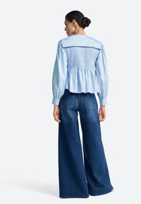 Blusa azul claro con espalda fruncida, mangas largas y dobladillo ancho, combinada con vaqueros de pierna ancha y talle alto en azul oscuro.