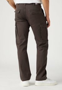 Mies, joka kantaa tummanruskeita cargo-housuja ja valkoisia tenniskenkiä, seisoo kädet sivuilla yksinkertaista taustaa vasten.