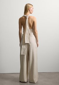 Beige Leinen-Overall mit einem Halterneck, der am Rücken gebunden ist, mit weitem Bein und einem offenen Rückendesign. Glatte Textur und minimalistisches Design.