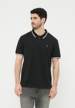 Άνδρας με σκούρα μαλλιά που φοράει μαύρο πόλο πουκάμισο με λευκή λεπτομέρεια και γκρι τζιν, στέκεται μπροστά σε απλό λευκό φόντο, κοιτώντας προς τα αριστερά.