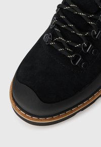 Schwarze Wildleder-Wanderstiefel mit schwarz-weißen Schnürsenkeln, Metallösen, Gummispitzen und mehrschichtiger braun-schwarzer Sohle.