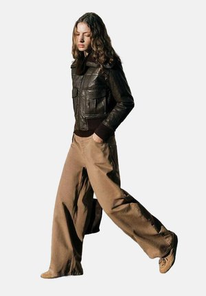 Brown leather jacket with ribbed cuffs, paired with wide khaki trousers and brown lace-up shoes. Model walking against a plain background.
