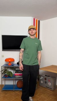 Νεαρός άνδρας με τατουάζ που φοράει πράσινο πουκάμισο και καφέ καπέλο στέκεται σε ένα δωμάτιο με τηλεόραση, πορτοκαλί λάμπα, μπάλα του μπάσκετ και μεταλλική βαλίτσα με αυτοκόλλητα.