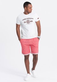 T-shirt blanc avec impression graphique rouge associé à un short rouge chiné, avec des ourlets roulés. Des baskets blanches complètent l'ensemble.