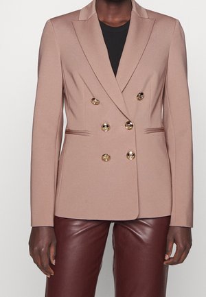 Beiger doppelt geknöpfter Blazer mit goldenen Knöpfen, getragen über einem schwarzen Top, kombiniert mit dunkelbraunen Lederhosen.