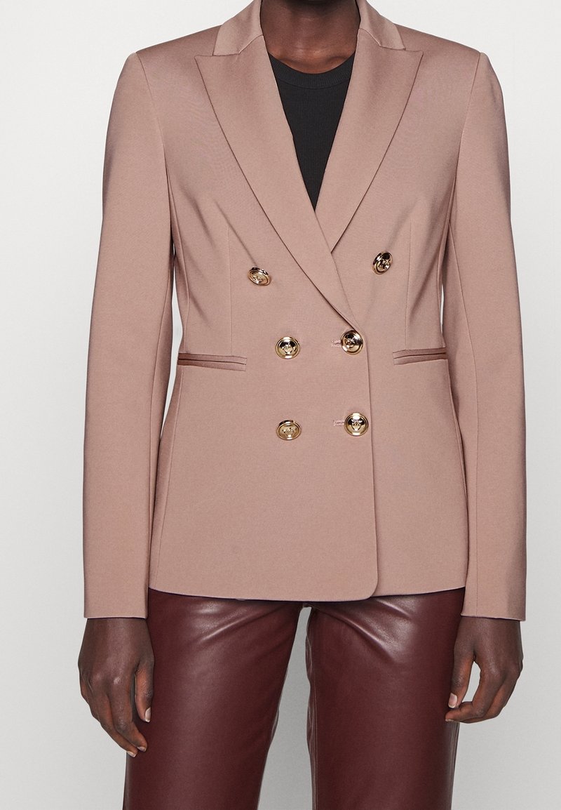 Blazer beige croisé avec des boutons dorés porté sur un top noir, associé à un pantalon en cuir marron foncé.