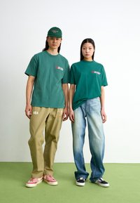 Tricouri din bumbac verde cu logo, pantaloni khaki cu nasturi și logo NBA, și adidași; unul roz, unul bleumarin. Design minimalist, croi lejer.