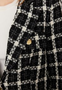 Kuvioitu mustavalkoinen tarkka blazeri, jossa on teksturoitu, kudottu kangas. Varustettu kultaisella napilla ja rakenteellisesti muotoilluilla kauluksilla. Kuvio luo ruudullisen efektin.