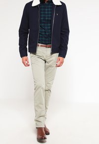 Giacca blu navy con zip e colletto in pelliccia ecologica, indossata sopra una camicia a quadri verde e blu navy, abbinata a pantaloni beige chiaro e stivali marroni.