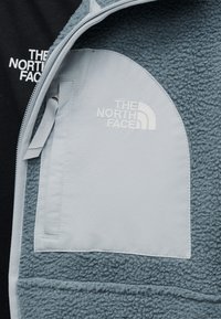 Ανοιχτόμπλε φλις μπουφάν με λευκή τσέπη στο στήθος με φερμουάρ και κεντημένο λογότυπο "The North Face" στην τσέπη και μαύρη εσωτερική επένδυση.