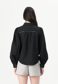 Svart knapp-up bluse med puffede lange ærmer og broderte detaljer langs sømmene, parret med lysegrå shorts.