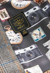 Stoff mit Vintage-Kameras, Uhren und Briefmarken in gedämpften Farben. Ein braun-blaues Karomuster ist auf der linken Seite sichtbar. Etiketten sind enthalten.