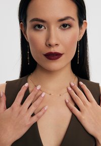 Złota biżuteria z wisiorkami w kształcie kolczyków i delikatnym naszyjnikiem z koralikami. Modelka ma długie ciemne włosy i jasnoróżowy lakier do paznokci.