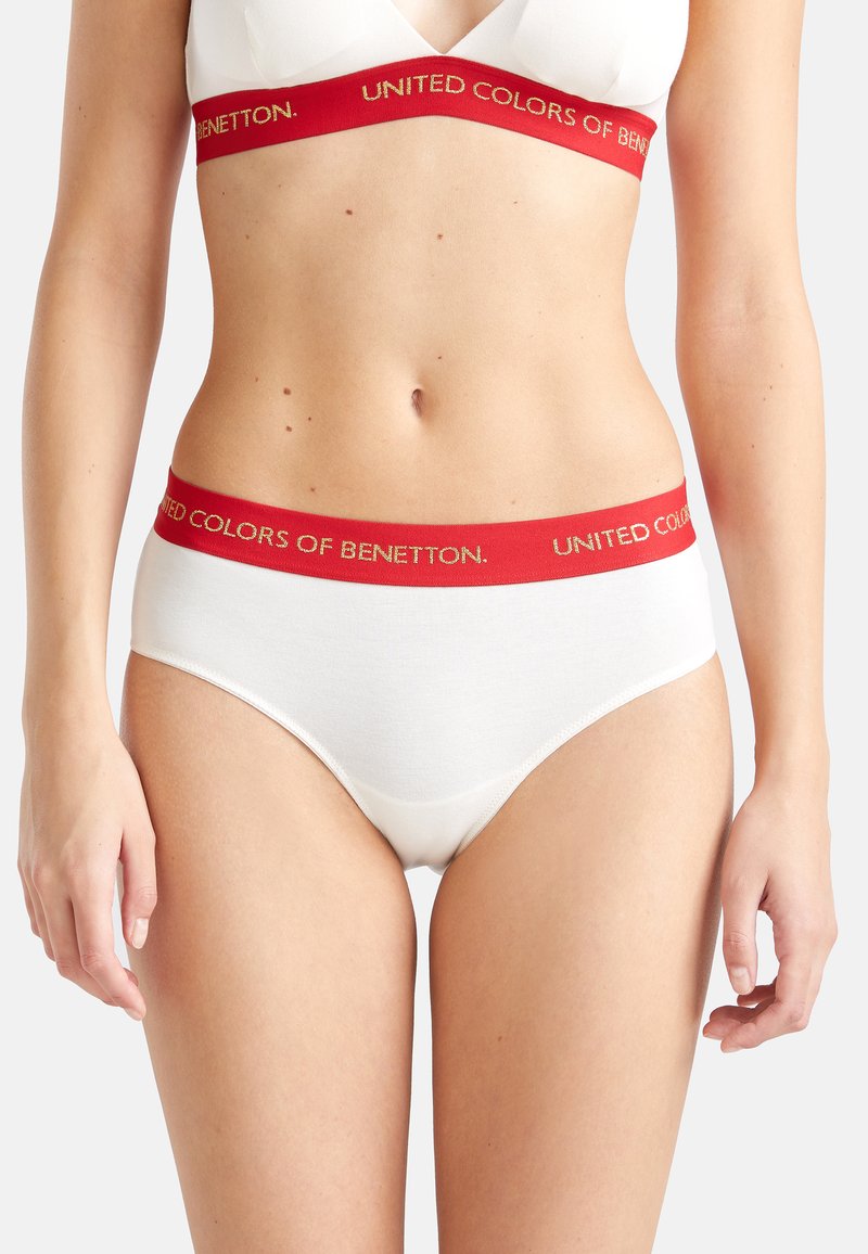 Donna che indossa un set di biancheria intima bianca con elastici rossi recanti il testo "United Colors of Benetton", mostrata dalla parte centrale del petto fino alle cosce superiori.