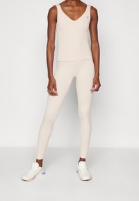 Persona con un top sin mangas de canalé beige claro y leggings a juego, llevando zapatillas blancas con acentos en púrpura y rosa.
