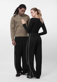 Barna csíkos pulóvert és fekete nadrágot viselő férfi áll egy fekete felsőt és fekete, fehér csíkos nadrágot viselő nő mellett, mindketten láncokat viselnek.