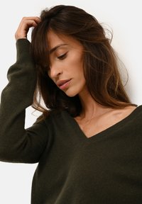 Tmavě zelený svetr s výstřihem do V vyrobený z měkké, strukturované tkaniny, s dlouhými rukávy a uvolněným střihem. Detailní pohled na výstřih.