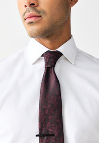 Burgunderroter Seidenschal mit einem strukturierten Blumenmuster, begleitet von einem schlanken Design und einem schwarzen Krawattenklammer, kombiniert mit einem weißen Hemd.