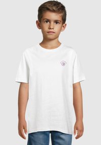Camiseta de algodón blanca con mangas cortas y escote redondo, que presenta un pequeño gráfico púrpura en el lado izquierdo del pecho. Combinada con jeans azules.