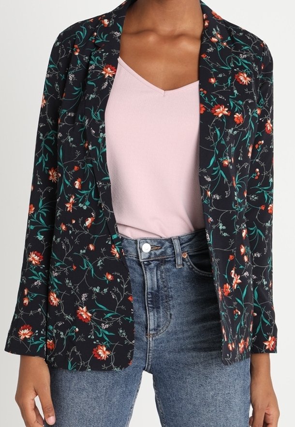 Femme portant un blazer noir à motifs floraux sur un haut rose clair, associé à un jean bleu taille haute, vue des épaules jusqu'aux hanches.