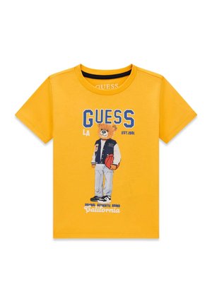 T-shirt jaune à manches courtes avec un ours en peluche de dessin animé tenant un ballon de basket, portant une veste universitaire et des baskets, avec le texte « GUESS » au-dessus.