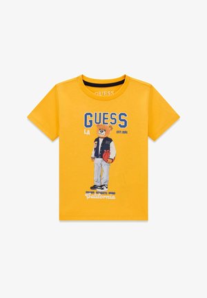T-shirt jaune à manches courtes avec un ours en peluche de dessin animé tenant un ballon de basket, portant une veste universitaire et des baskets, avec le texte « GUESS » au-dessus.