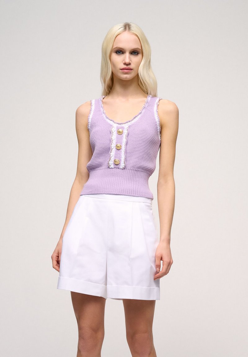 Levandulový pletený top s ribbed texturou, se třemi zlatými knoflíky a bílým třásněním, v kombinaci s bílými skládacími šortkami.