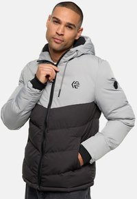 Chaqueta acolchada con cremallera, que tiene una parte superior gris y un fondo negro, con capucha, detalles en negro y un logotipo en el pecho. Acabado texturizado.