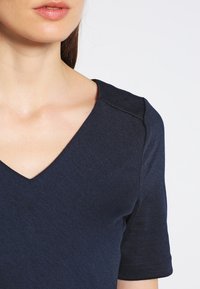 Haut bleu marine à col en V en tissu texturé, avec des manches courtes et un design épuré et moderne, agrémenté de détails de coutures visibles.