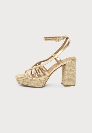 Sandale dorée à talons hauts avec brides, boucle à la cheville, plateforme tressée et talon bloc sur fond blanc.